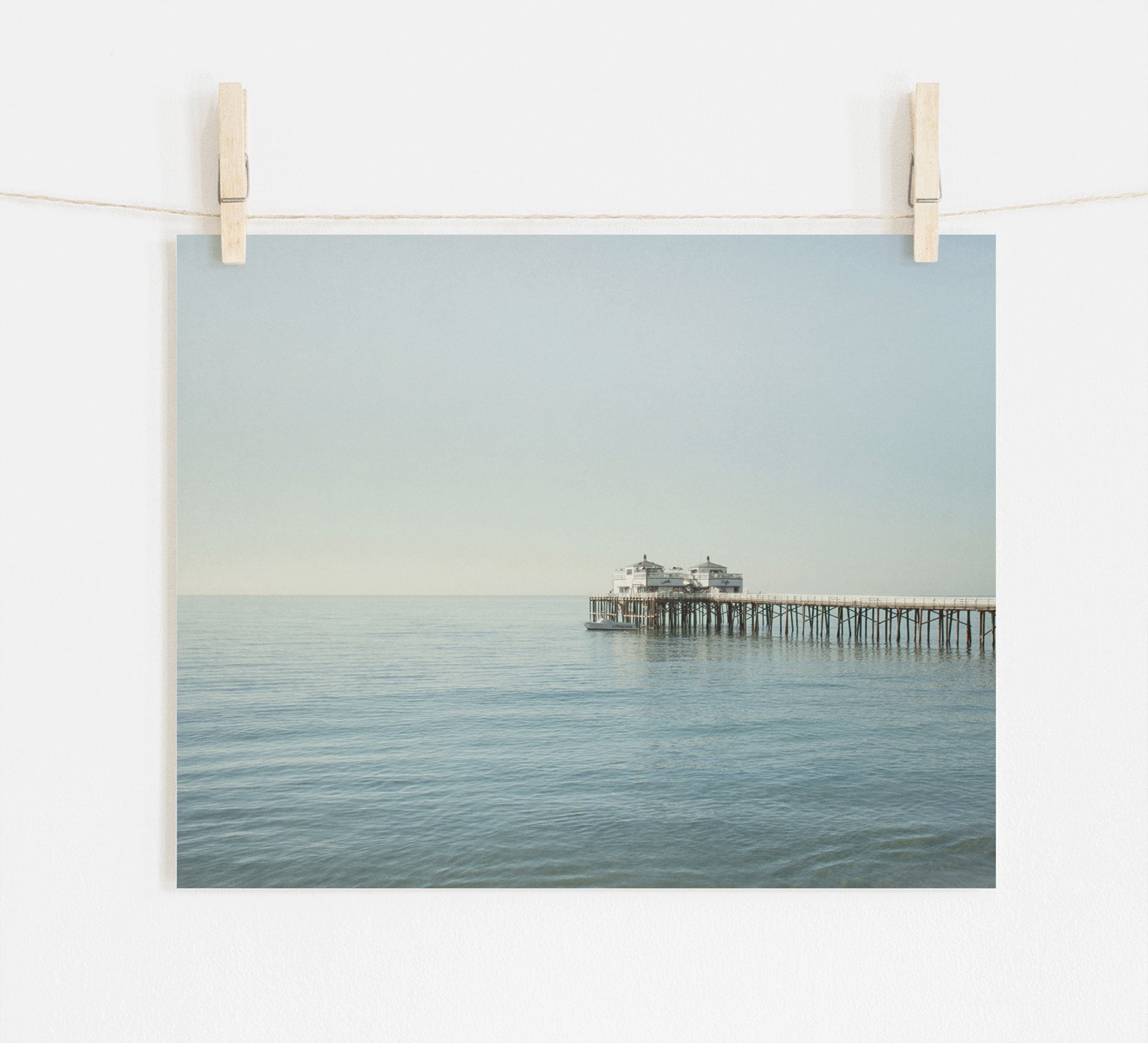 A tranquil photograph of Malibu Pier extending into a calm sea, pinned up against a white wall by two wooden clothespins on a string. Coastal Print of Malibu Pier in California &#39;All Calm in Malibu&#39; by Offley Green.