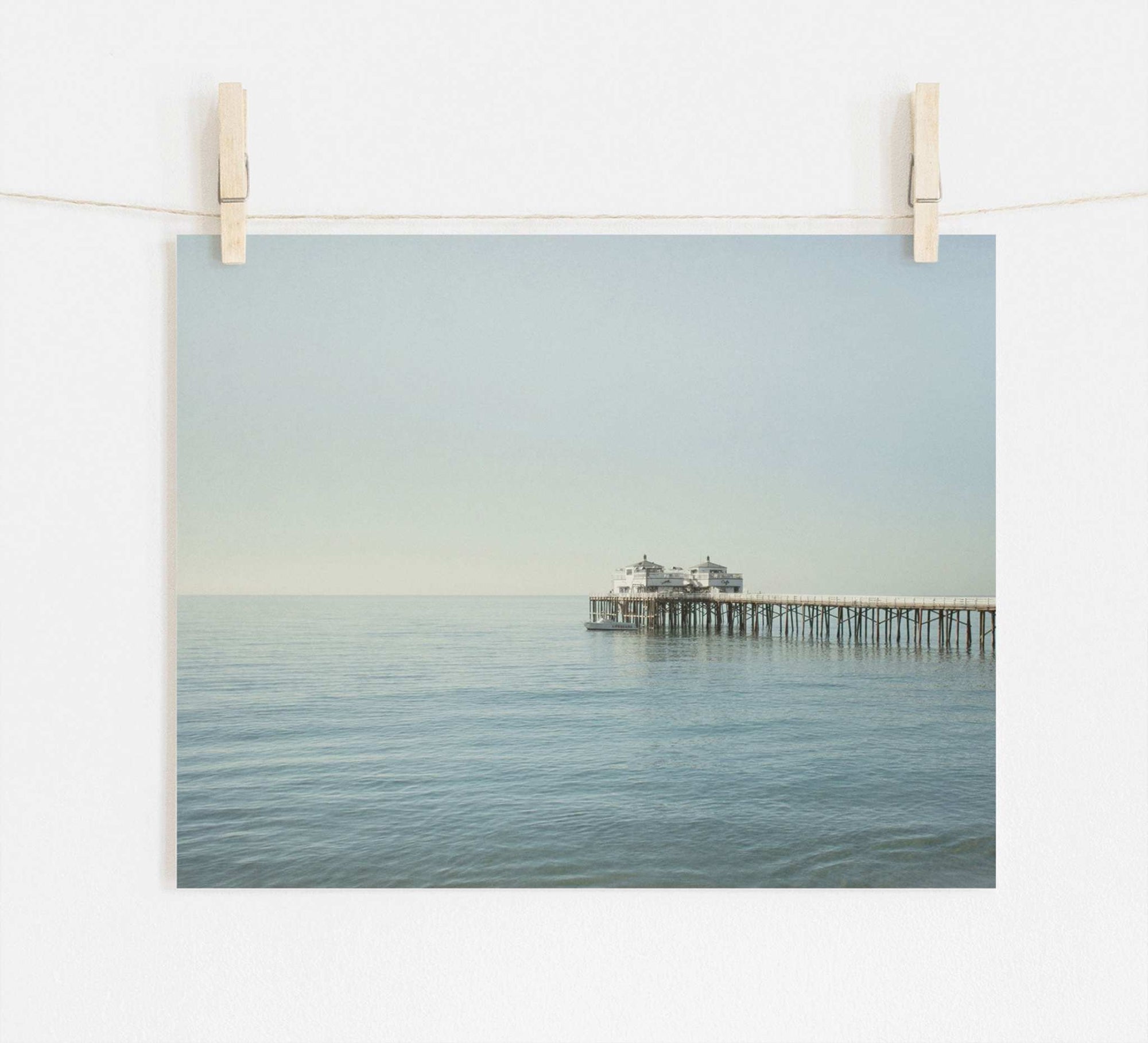 A tranquil photograph of Malibu Pier extending into a calm sea, pinned up against a white wall by two wooden clothespins on a string. Coastal Print of Malibu Pier in California 'All Calm in Malibu' by Offley Green.