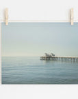 A tranquil photograph of Malibu Pier extending into a calm sea, pinned up against a white wall by two wooden clothespins on a string. Coastal Print of Malibu Pier in California 'All Calm in Malibu' by Offley Green.