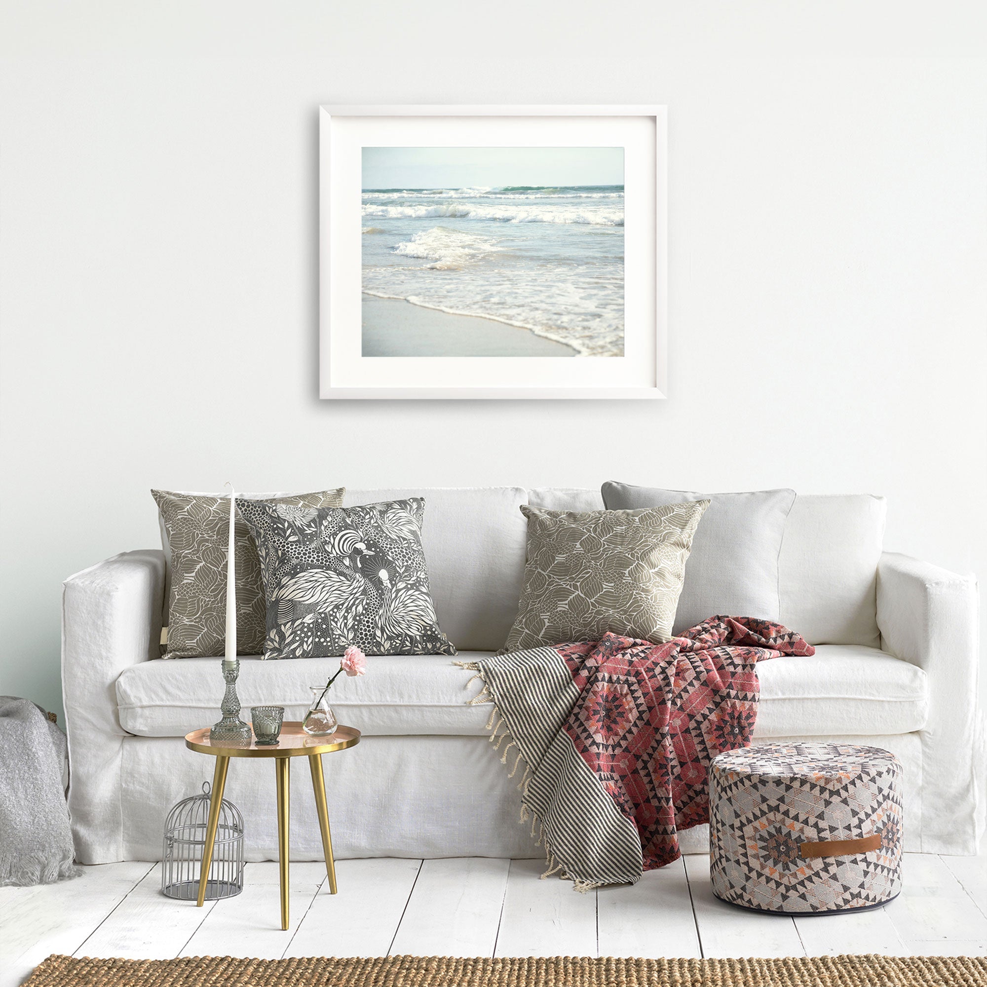 A cozy living room setup with a white Offley Green sofa adorned with the Coastal Beach Print in California 'Surf and Sun' patterned cushions, a small gold side table, a wicker ottoman with a pink flower on top, and a gray cage on the floor.