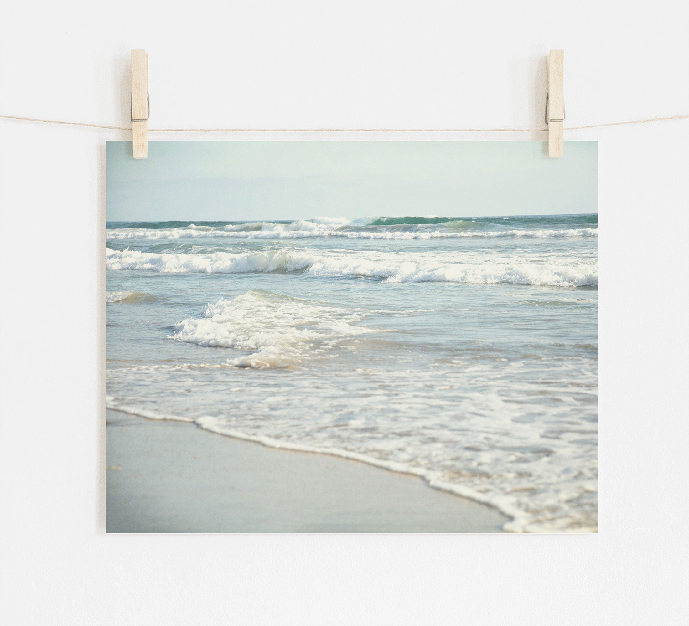 A photograph of Carlsbad State Beach showing waves and shoreline, pinned by wooden clothespins on a string against a white wall. 
Product Name: Offley Green Coastal Beach Print in California 'Surf and Sun'