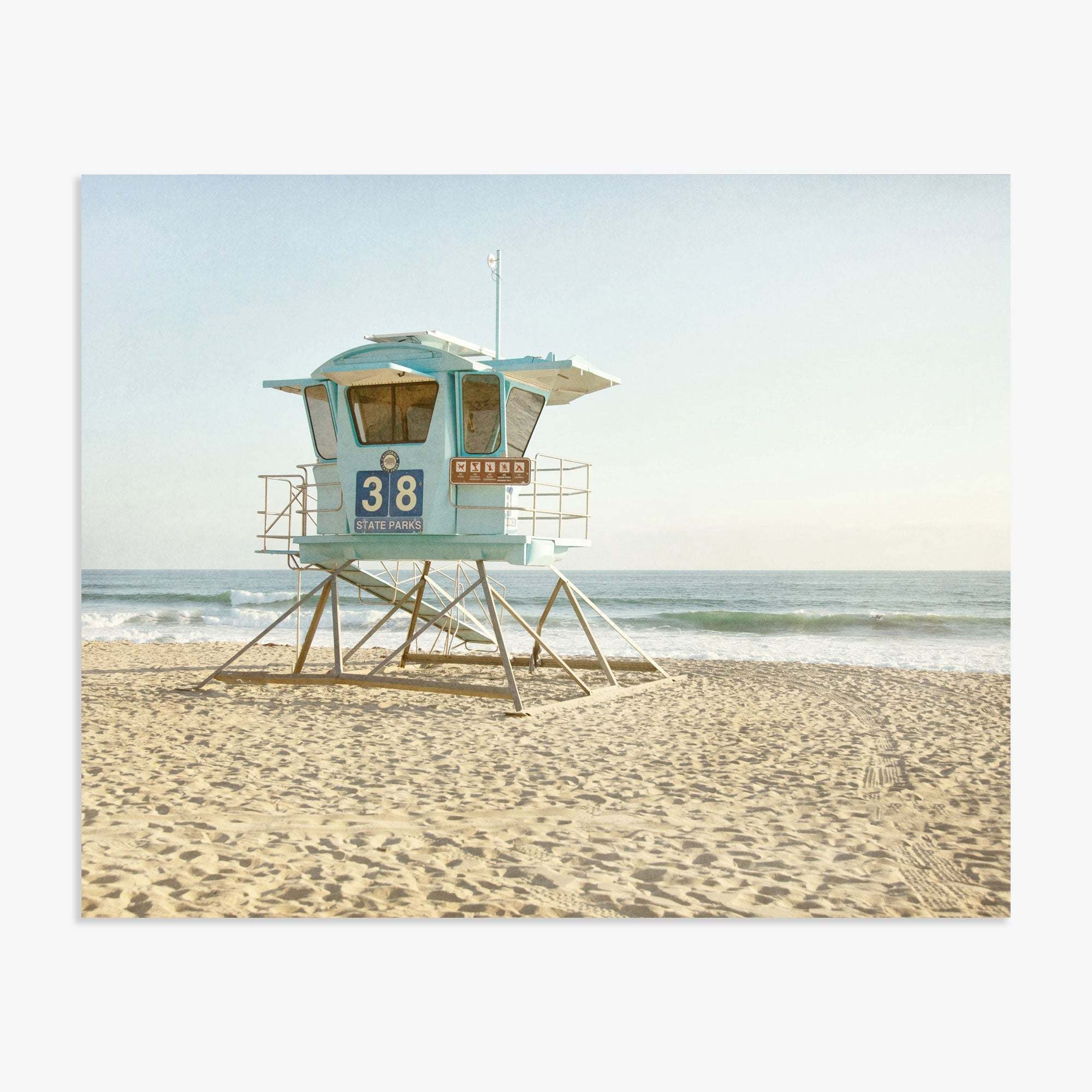 A picturesque scene of a lifeguard station, number 38, on a sandy beach with the ocean in the background under a clear sky, captured on Offley Green&#39;s California Coastal Print, &#39;Carlsbad Lifeguard Tower.&#39;