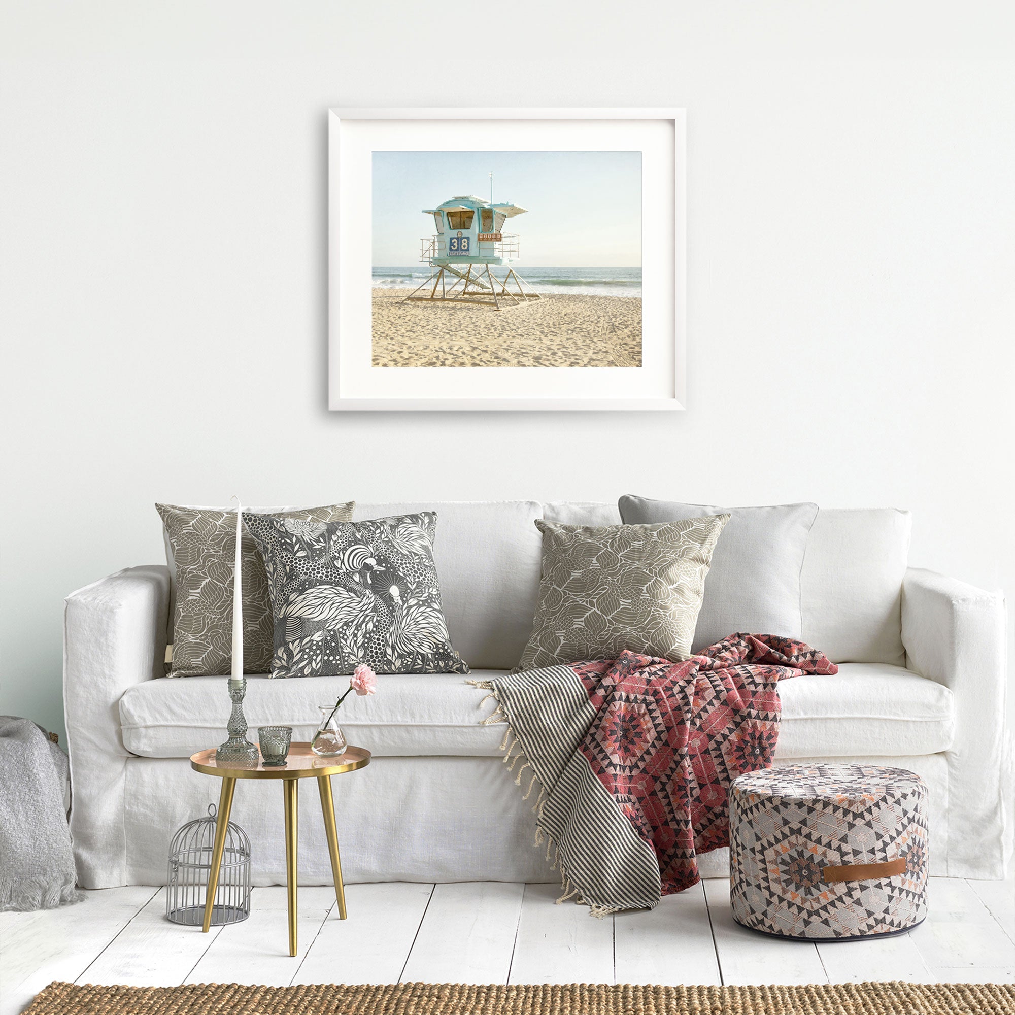 A cozy living room featuring a white sofa adorned with patterned cushions, a small table with decor, a wicker ottoman, and a framed print of the California Coastal Print, &#39;Carlsbad Lifeguard Tower&#39; by Offley Green hanging on the wall.