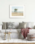 A cozy living room featuring a white sofa adorned with patterned cushions, a small table with decor, a wicker ottoman, and a framed print of the California Coastal Print, 'Carlsbad Lifeguard Tower' by Offley Green hanging on the wall.