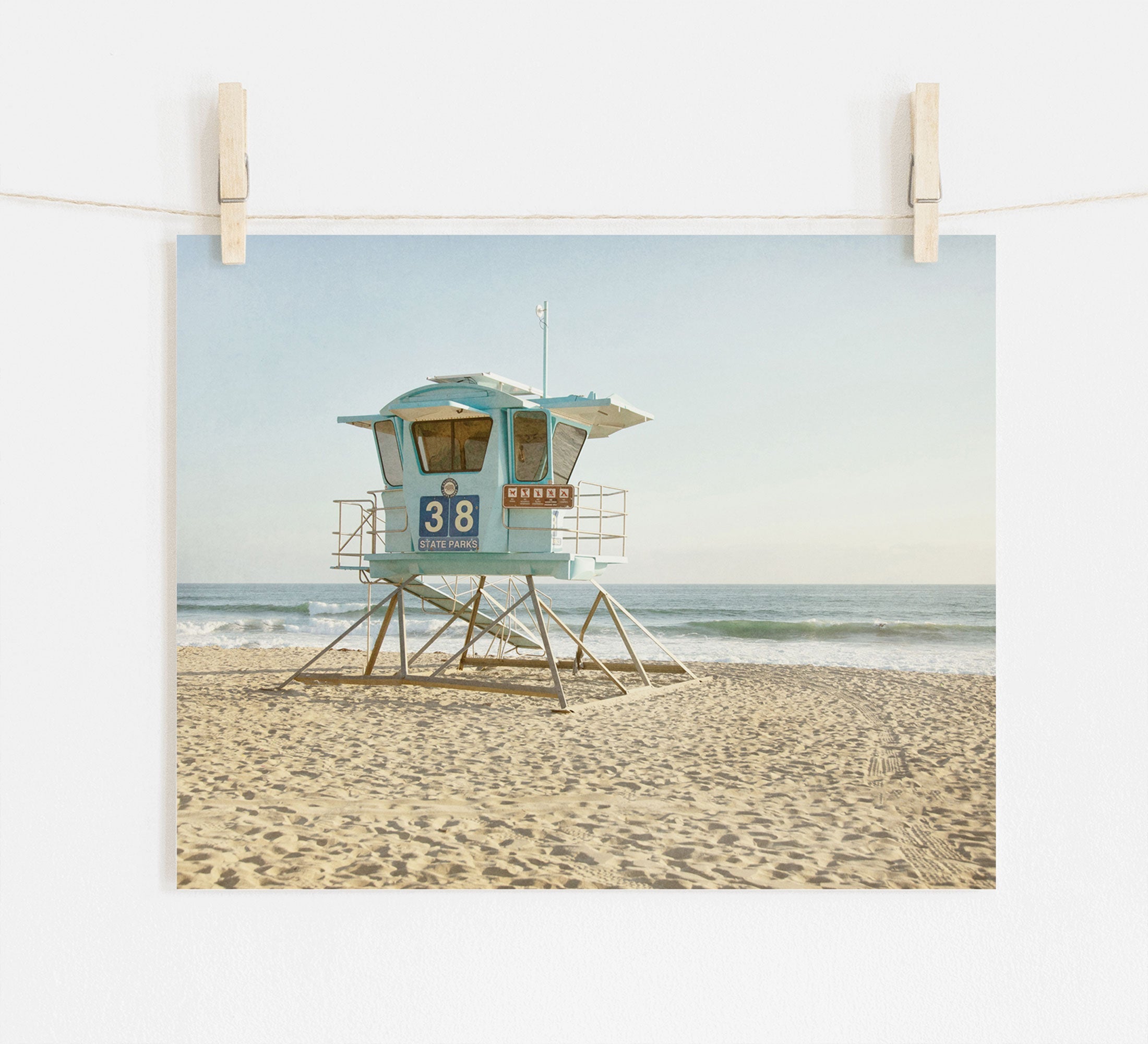 A photograph of a light blue lifeguard tower numbered 38 on a sandy beach, printed on archival photographic paper, displayed on a beige wall, held by two wooden pegs. Waves gently lap the Offley Green California Coastal Print, &#39;Carlsbad Lifeguard Tower&#39;.