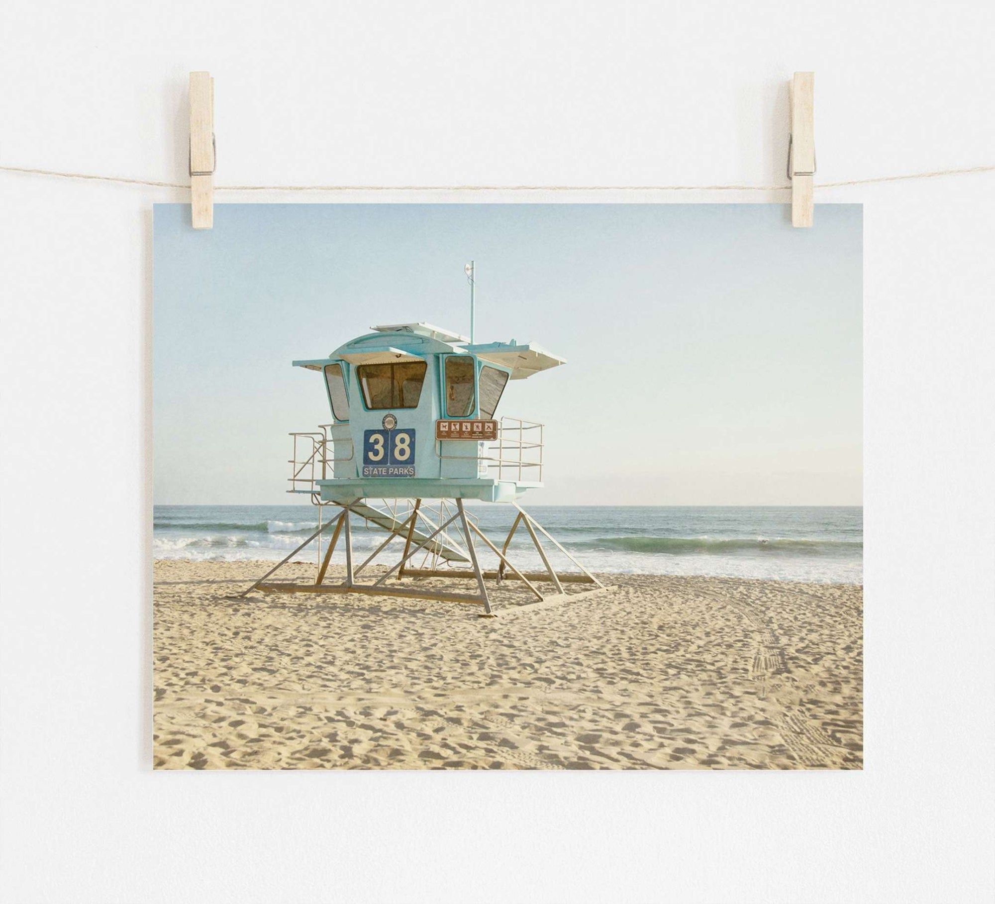 A photograph of a light blue lifeguard tower numbered 38 on a sandy beach, printed on archival photographic paper, displayed on a beige wall, held by two wooden pegs. Waves gently lap the Offley Green California Coastal Print, 'Carlsbad Lifeguard Tower'.