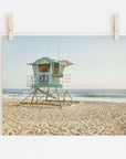 A photograph of a light blue lifeguard tower numbered 38 on a sandy beach, printed on archival photographic paper, displayed on a beige wall, held by two wooden pegs. Waves gently lap the Offley Green California Coastal Print, 'Carlsbad Lifeguard Tower'.