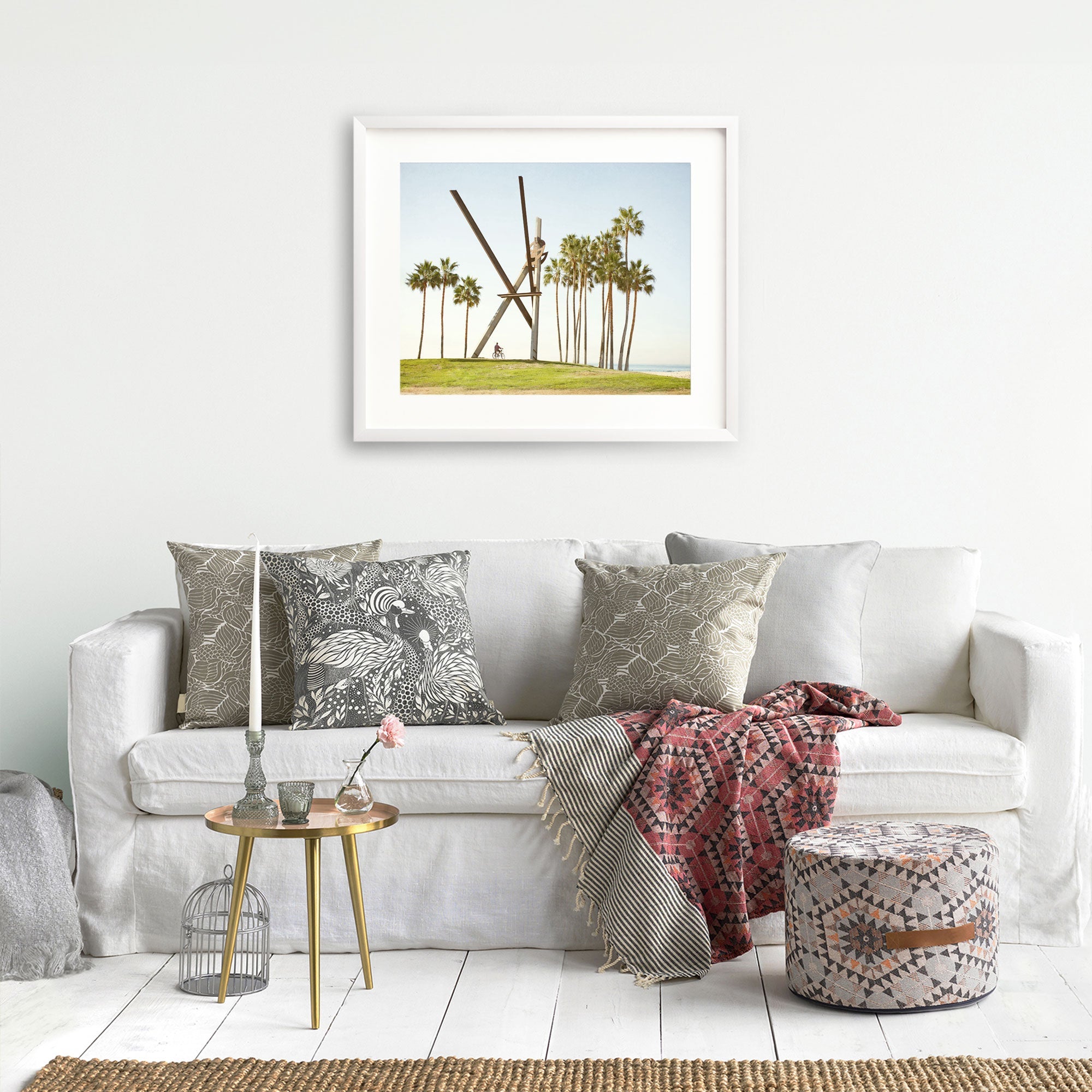 A cozy living room featuring a white sofa with decorative pillows, a red patterned throw blanket, a small gold side table, and an unframed Offley Green Venice Beach Landmark Sculpture, 'V is for Venice' photography hanging above the couch.