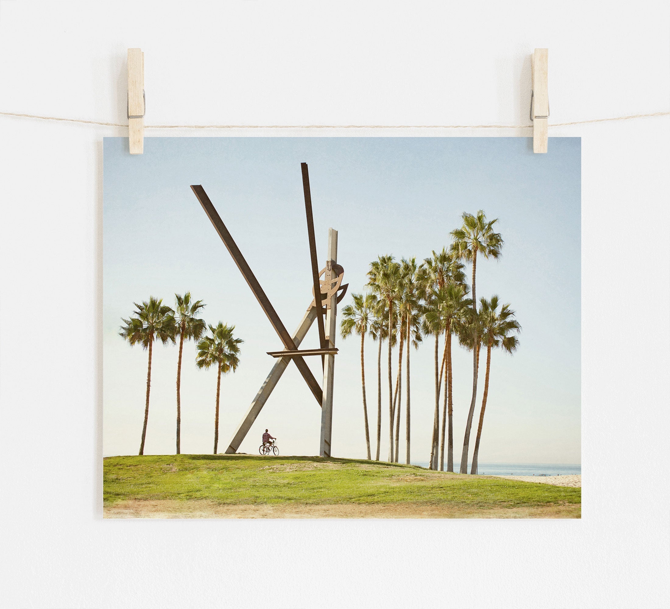 A photograph of the Venice Beach Landmark Sculpture, 'V is for Venice', by Offley Green, surrounded by tall palm trees, printed on archival photographic paper, hangs on a white wall displayed with wooden clips on a string. In the background, a person on a.