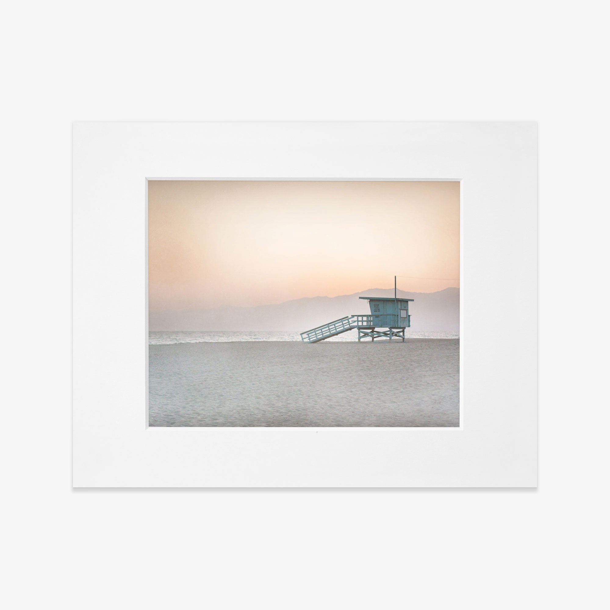 A serene beach scene featuring a Pink Coastal Print, &#39;Lifeguard Tower&#39; centered on a deserted shore with distant mountains, under a soft gradient sky from peach to pale blue, captured on archival photographic paper with a non glossy lustre by Offley Green.