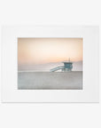 A serene beach scene featuring a Pink Coastal Print, 'Lifeguard Tower' centered on a deserted shore with distant mountains, under a soft gradient sky from peach to pale blue, captured on archival photographic paper with a non glossy lustre by Offley Green.