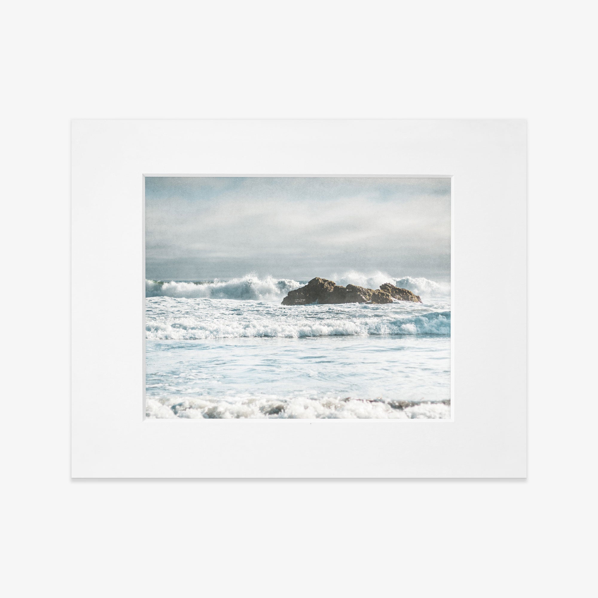 A framed photograph depicts a moody seascape at Pfeiffer Beach, with waves crashing against rocky formations in the distance. The sky above is partially cloudy, adding a soft, calming atmosphere to the image. The white frame contrasts with the vibrant blues and whites of the ocean, printed on archival photographic paper. This piece is known as Seascape Coastal Art, &#39;Surf and Rocks&#39; by Offley Green.