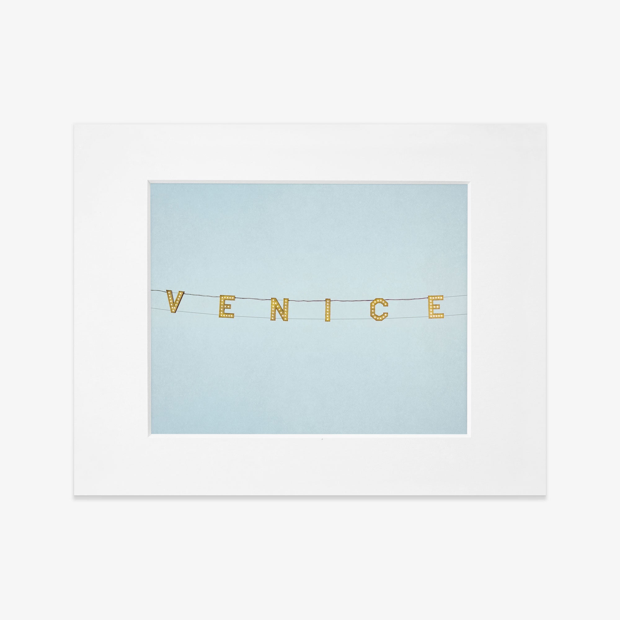 A framed photograph featuring the Venice Beach Sign Print, &#39;Blue Venice&#39; spelled out on small, gold, circular pieces strung together against a soft blue background, printed on archival photographic paper by Offley Green.