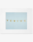 A framed photograph featuring the Venice Beach Sign Print, 'Blue Venice' spelled out on small, gold, circular pieces strung together against a soft blue background, printed on archival photographic paper by Offley Green.