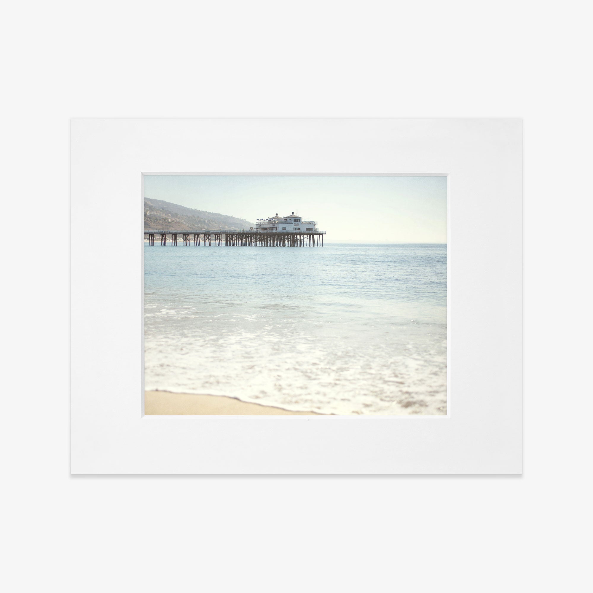A serene beachscape featuring gentle waves washing ashore, with the Offley Green California Beach Print, &#39;Malibu Pier&#39; extending into the sea under a clear blue sky, framed as a polaroid photo.