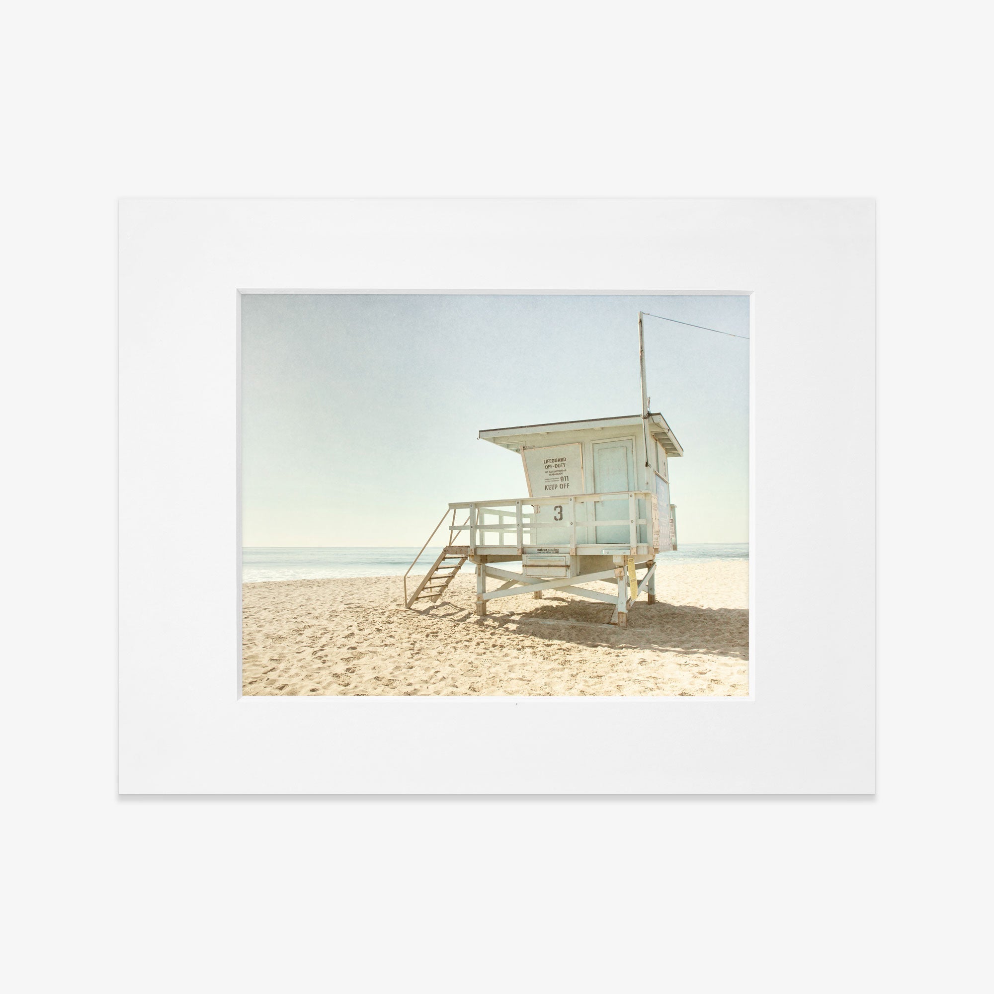 A vintage-style image of a white lifeguard tower in Malibu, number 3, situated on a sandy beach against a clear sky. The scene is tranquil with no people visible, emphasizing solitude. This is Offley Green&#39;s California Summer Beach Art, &#39;Malibu Lifeguard Tower&#39;.