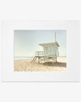 A vintage-style image of a white lifeguard tower in Malibu, number 3, situated on a sandy beach against a clear sky. The scene is tranquil with no people visible, emphasizing solitude. This is Offley Green's California Summer Beach Art, 'Malibu Lifeguard Tower'.