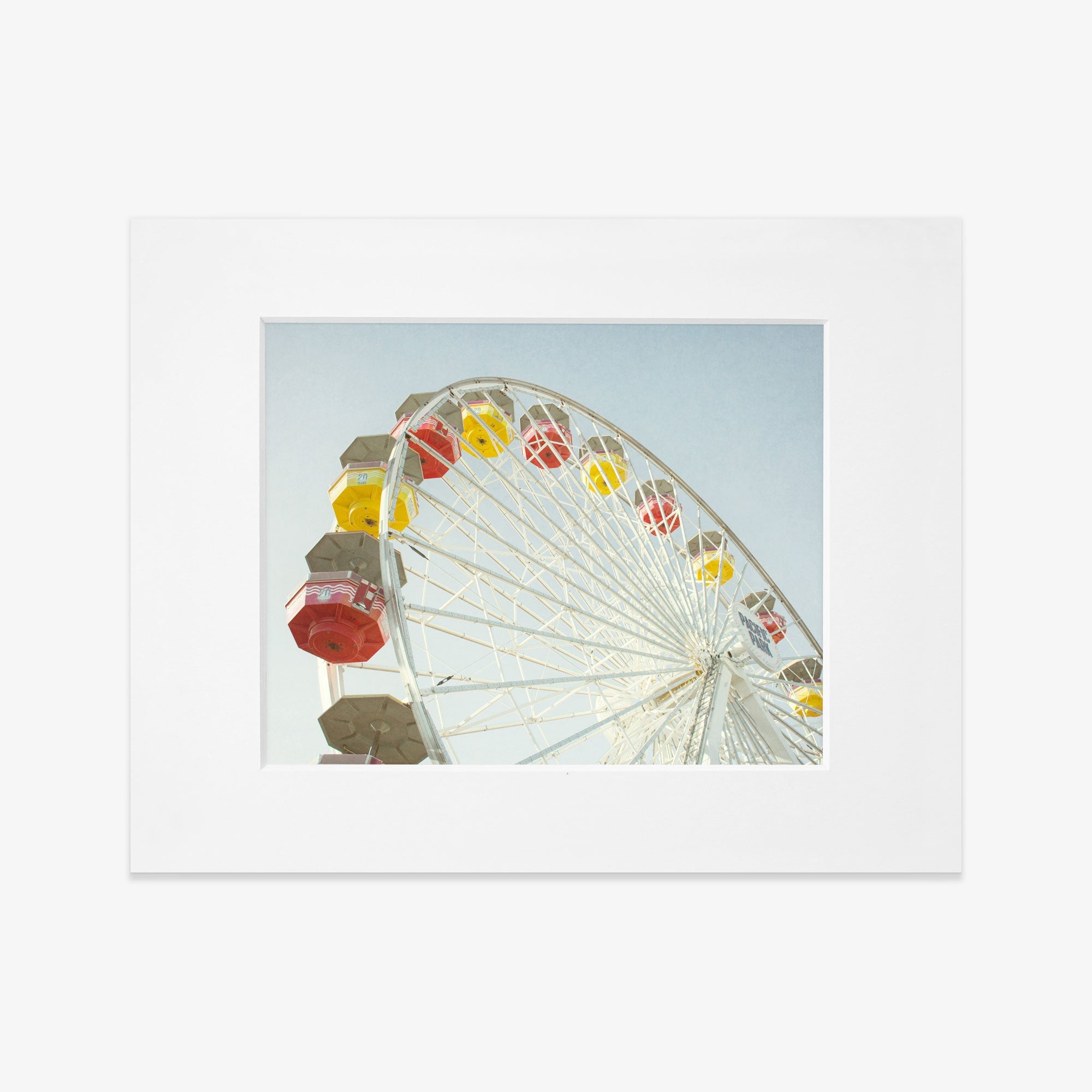 A colorful Santa Monica Ferris Wheel Print, &#39;Ferris Above&#39; at Santa Monica Pier set against a clear blue sky, framed in a simple white border by Offley Green.