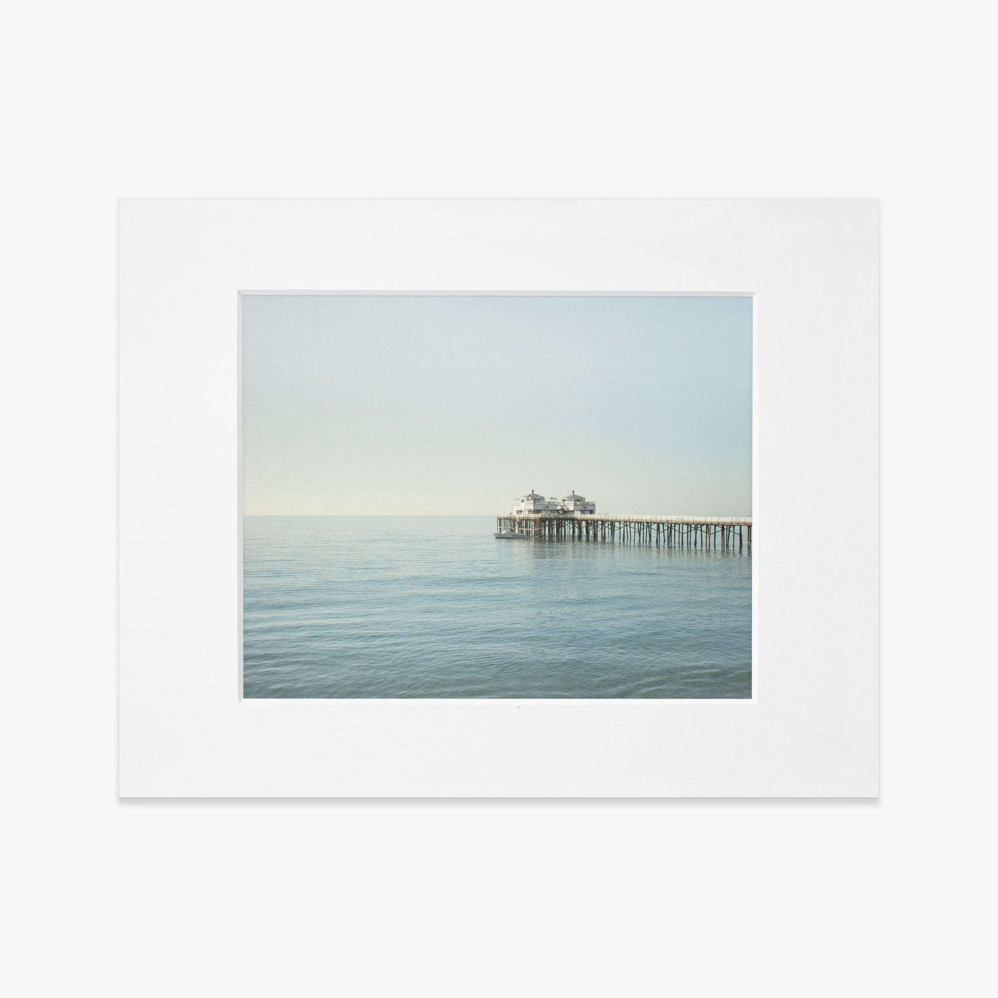 A serene image of Malibu Pier extending into a calm sea under a clear sky, framed in white, capturing a peaceful maritime scene - Offley Green&#39;s &#39;All Calm in Malibu&#39; coastal print.