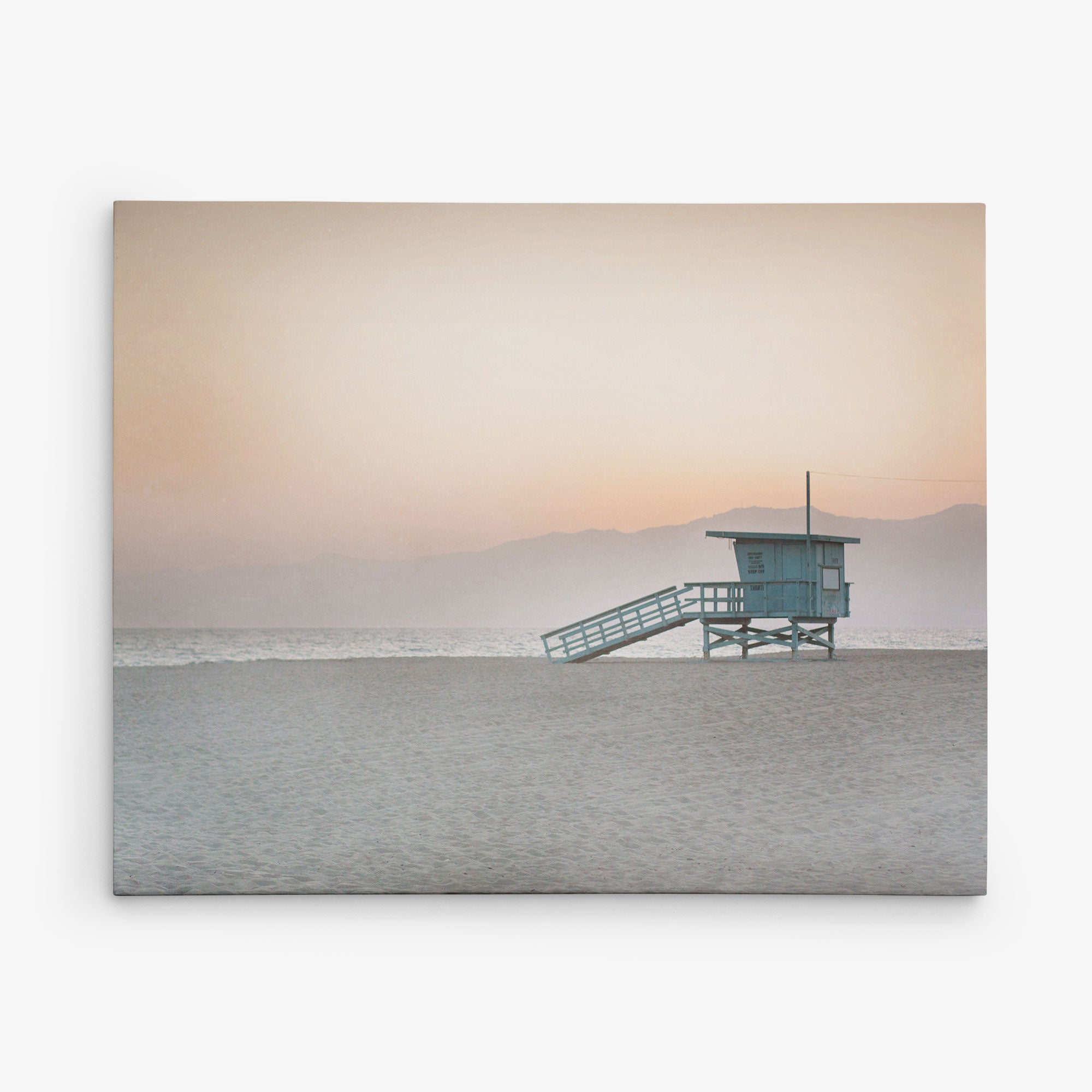 A serene beach scene at sunset featuring a solitary Offley Green lifeguard tower against a soft pink sky with marine layer mist and mountain silhouettes in the background.