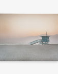 A serene beach scene at sunset featuring a solitary Offley Green lifeguard tower against a soft pink sky with marine layer mist and mountain silhouettes in the background.