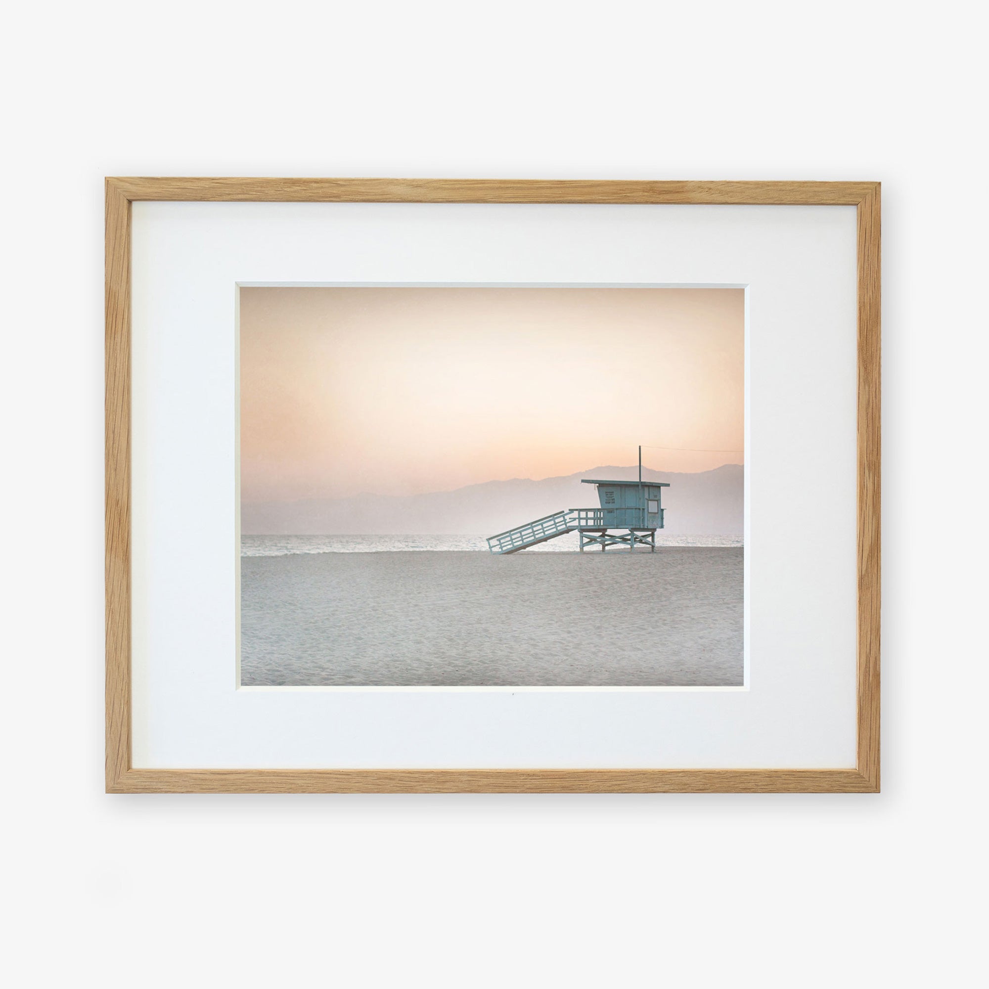 A framed photograph of a Pink Coastal Print, &#39;Lifeguard Tower&#39; by Offley Green, featuring a serene beach scene at sunrise with a solitary lifeguard station and misty mountains in the background, displayed on a white wall with a non-glossy lustre finish.