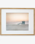 A framed photograph of a Pink Coastal Print, 'Lifeguard Tower' by Offley Green, featuring a serene beach scene at sunrise with a solitary lifeguard station and misty mountains in the background, displayed on a white wall with a non-glossy lustre finish.