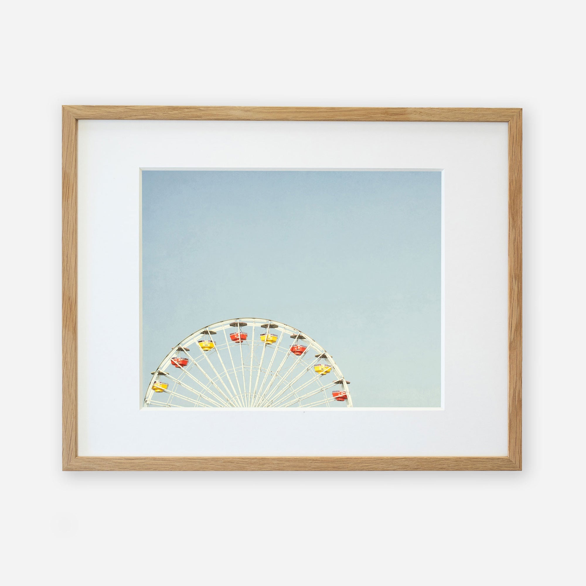 A framed photograph of Ferris Blue, the Santa Monica Pier ferris wheel, partially visible at the bottom, against a light blue sky, displayed in a simple wooden frame with a white mat by Offley Green.