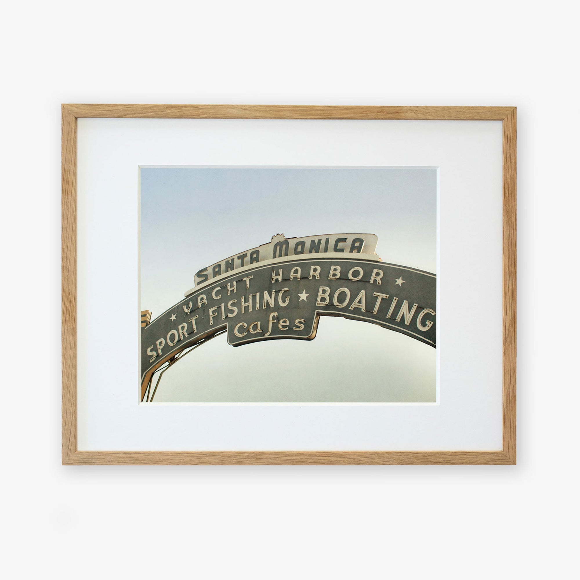 Framed archival photographic print of the Los Angeles California Print, 'Santa Monica Pier Blues' by Offley Green, featuring the iconic Santa Monica sign with text highlighting yacht harbor, sport fishing, boating, and cafes, displayed against a clear sky.