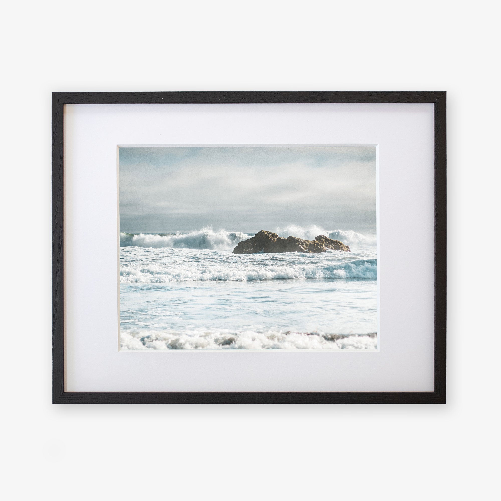 A framed photograph displays a serene ocean scene with waves crashing against rocks under a cloudy sky at Pfeiffer Beach. The moody seascape, printed on archival photographic paper, features a mix of blue and white hues, capturing the dynamic movement of the water and the tranquility of the seaside atmosphere. This is Offley Green&#39;s Seascape Coastal Art, &#39;Surf and Rocks&#39;.