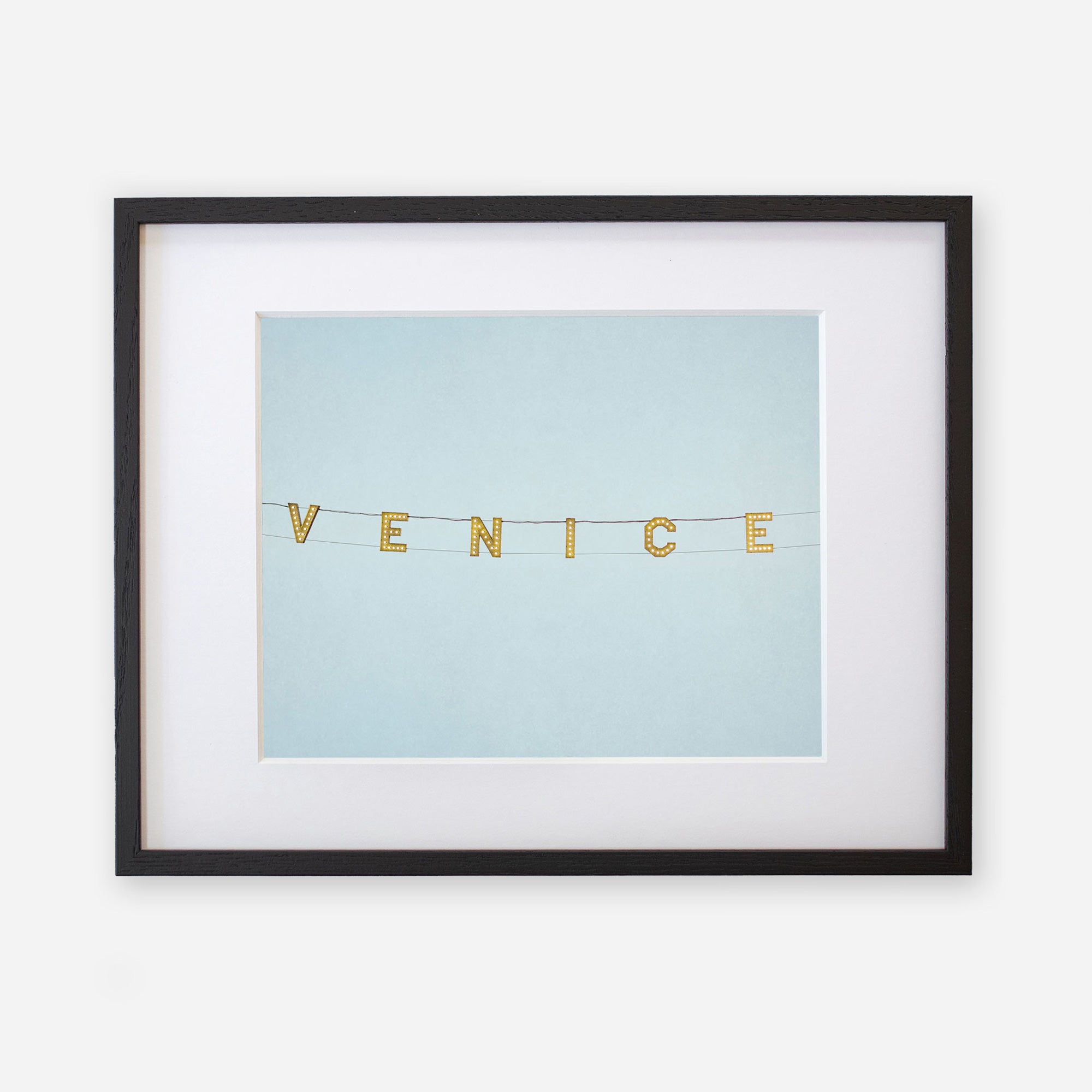 A framed artwork featuring the Venice Beach Sign Print, &#39;Blue Venice&#39; spelled out in gold letters on a pale blue background, printed on archival photographic paper, encased in a black frame with a white mat by Offley Green.