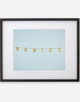 A framed artwork featuring the Venice Beach Sign Print, 'Blue Venice' spelled out in gold letters on a pale blue background, printed on archival photographic paper, encased in a black frame with a white mat by Offley Green.