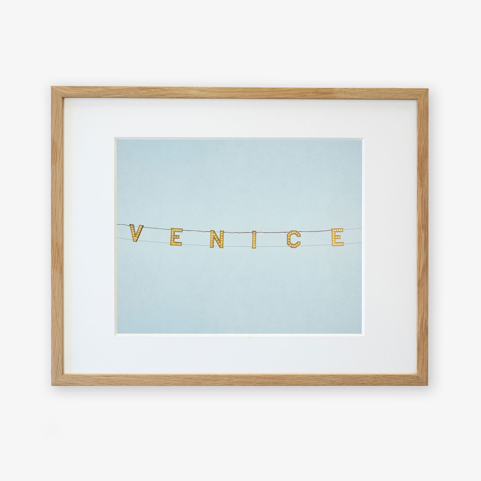 A framed wall art features the product &quot;Venice Beach Sign Print, &#39;Blue Venice&#39;&quot; spelled out on small square tiles that are strung together, displayed against a pale blue background on archival photographic paper by Offley Green.