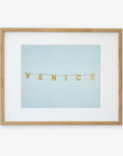 A framed wall art features the product "Venice Beach Sign Print, 'Blue Venice'" spelled out on small square tiles that are strung together, displayed against a pale blue background on archival photographic paper by Offley Green.