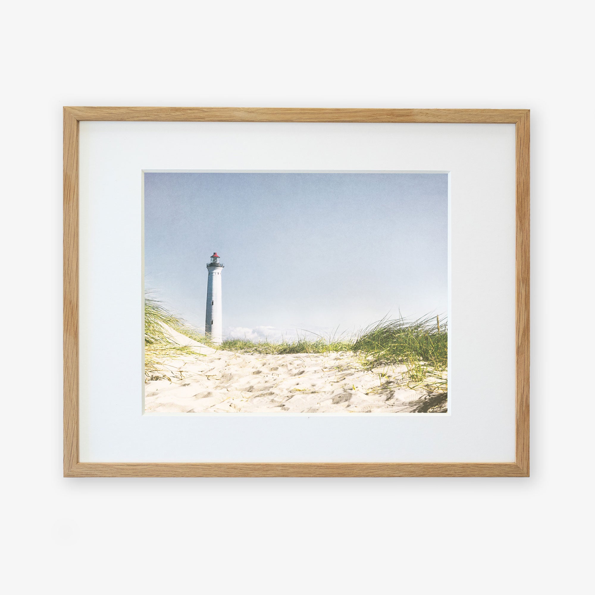 A framed Nautical Print, &#39;The Lighthouse&#39; by Offley Green of a lighthouse viewed from a sandy beach overgrown with tall grass, displayed against a white background. The lighthouse stands tall against a clear blue sky.