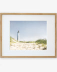 A framed Nautical Print, 'The Lighthouse' by Offley Green of a lighthouse viewed from a sandy beach overgrown with tall grass, displayed against a white background. The lighthouse stands tall against a clear blue sky.
