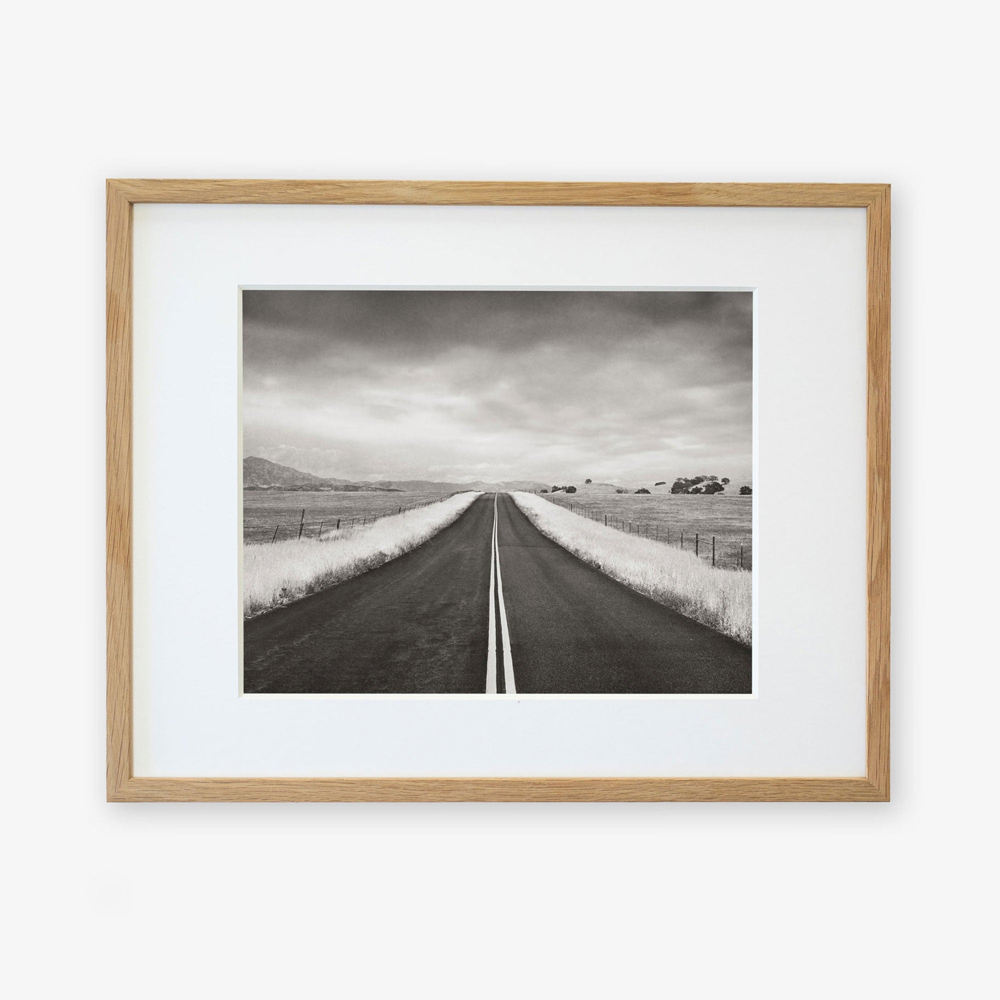 A framed black-and-white photograph depicting a straight road extending through a rural landscape under a cloudy sky, printed on archival photographic paper, displayed on a white background. This is the Offley Green Black and White Rural Landscape Art print entitled 'American Road Trip'.