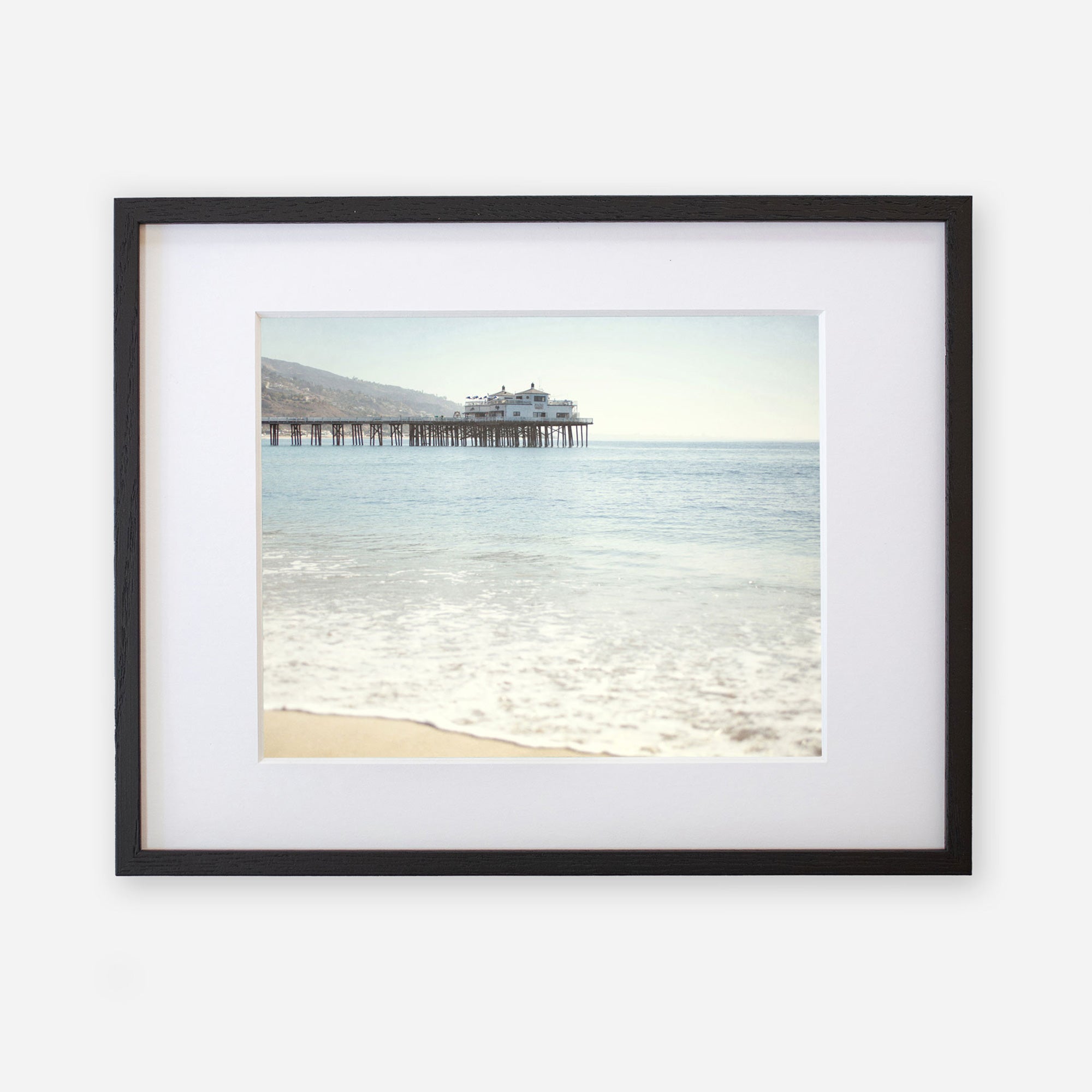 A framed photograph of a serene beach scene, showing gentle waves lapping against the shore with &#39;Malibu Pier&#39; extending into a calm sea under a clear sky by Offley Green.
