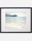 A framed photograph of a serene beach scene, showing gentle waves lapping against the shore with 'Malibu Pier' extending into a calm sea under a clear sky by Offley Green.