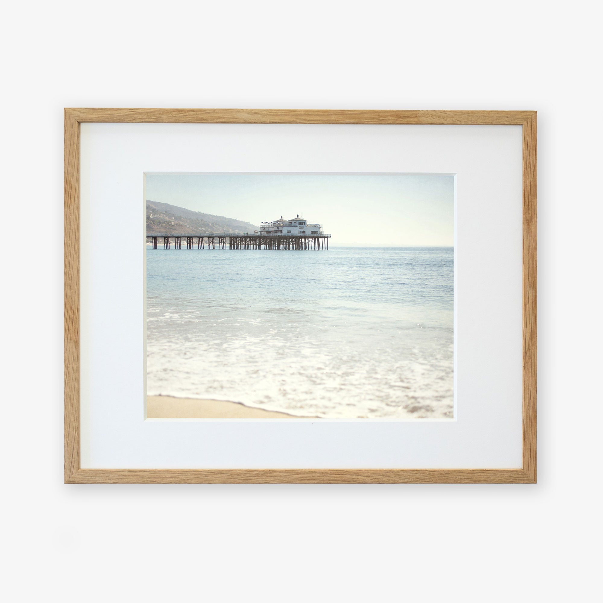 Framed photograph of a tranquil beach scene with gentle waves lapping the shore and California Beach Print, 'Malibu Pier' extending into the calm blue sea, all under a clear sky by Offley Green.