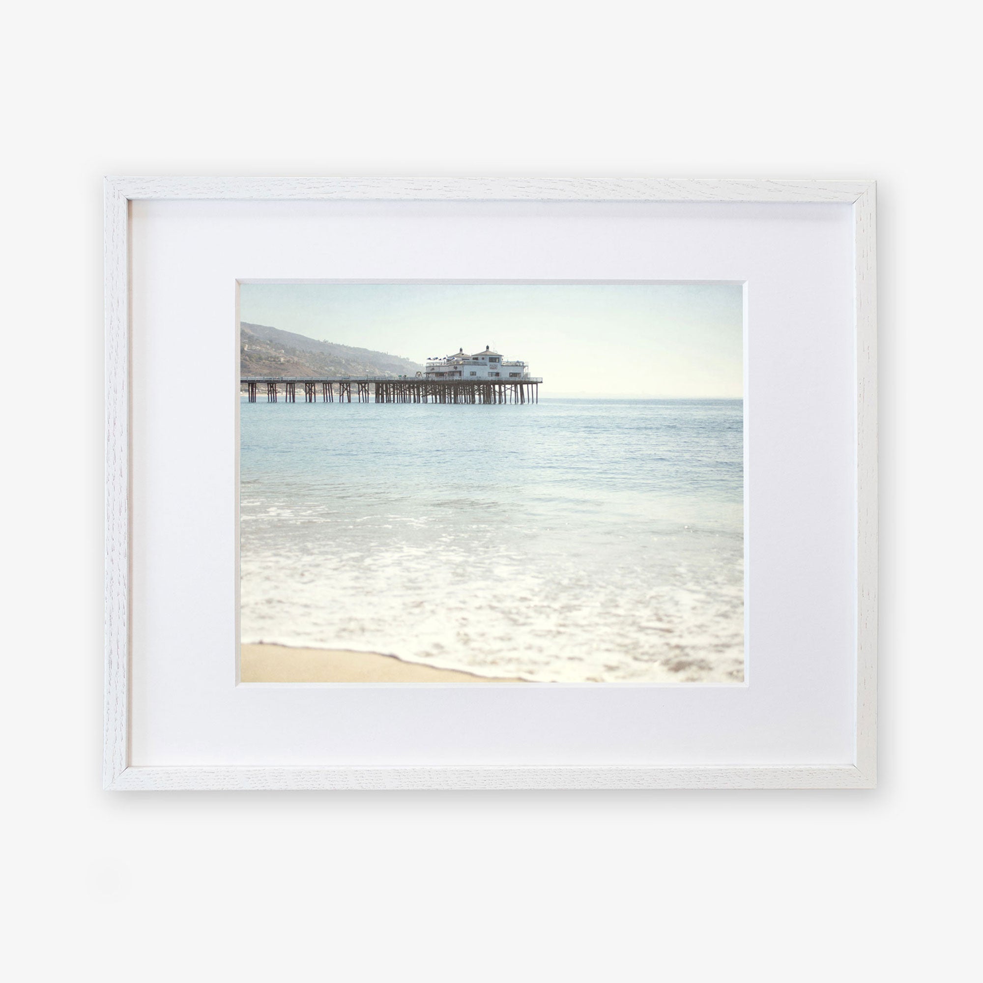 Framed photograph of a peaceful beach with gentle waves lapping at the shore, showcasing &#39;Malibu Pier&#39; extending into the calm blue ocean under a clear sky - Offley Green&#39;s California Beach Print.