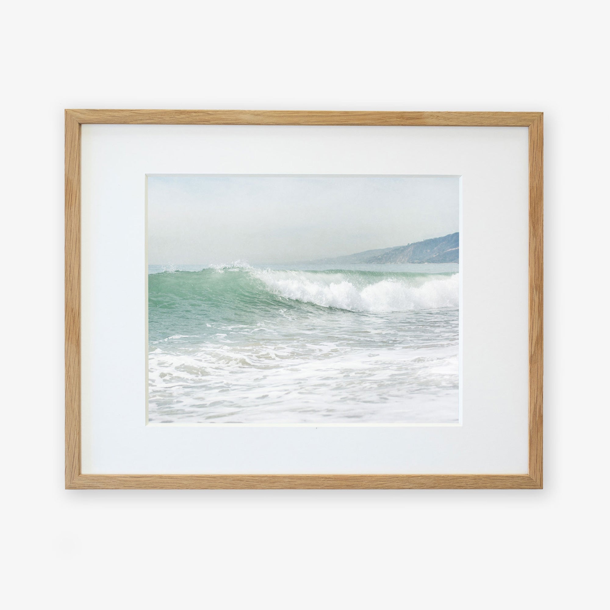 A framed photograph of a wave captured at the moment it breaks on a Southern California beach, with a foamy crest and misty backdrop, hung on a plain white wall. Offley Green's Coastal Print of a Breaking Wave 'Breaking Surf'