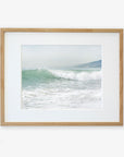 A framed photograph of a wave captured at the moment it breaks on a Southern California beach, with a foamy crest and misty backdrop, hung on a plain white wall. Offley Green's Coastal Print of a Breaking Wave 'Breaking Surf'