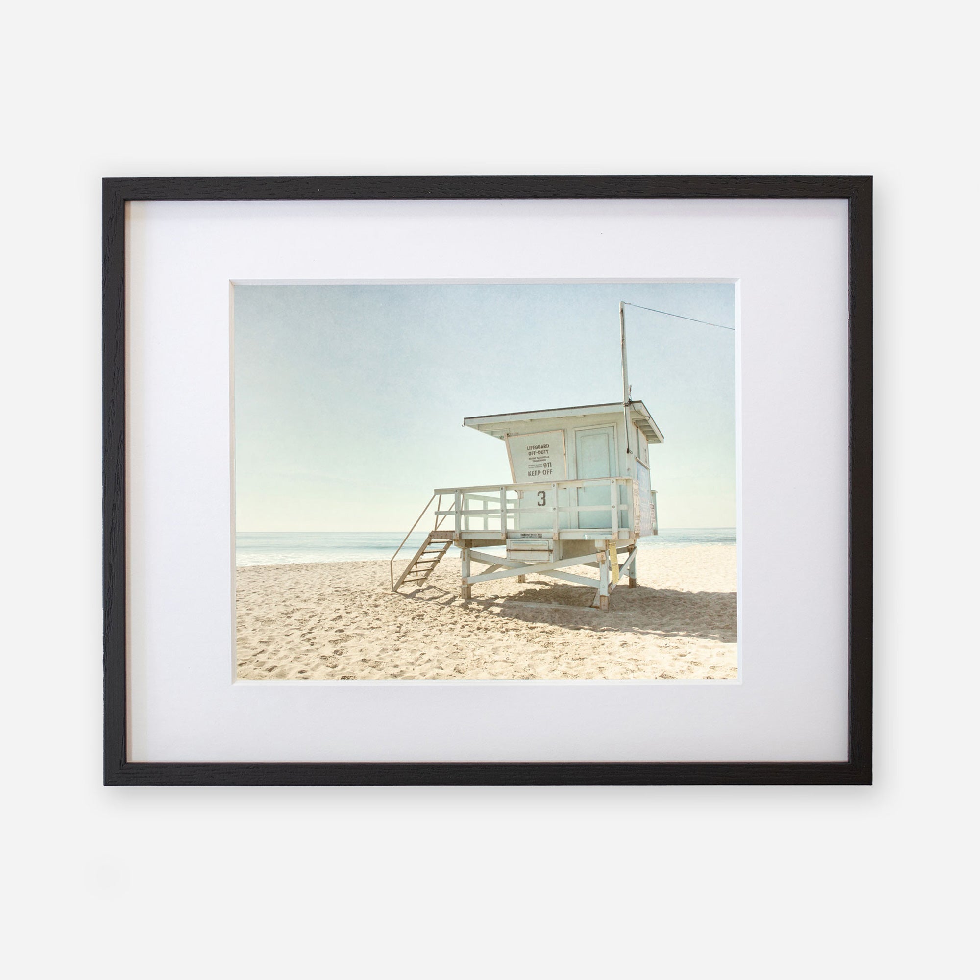 A framed Offley Green photograph depicts a lone lifeguard tower in Malibu on a sandy beach under a clear sky, creating a serene coastal scene.
