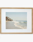 A framed photograph of a Malibu Beach House Print, 'Ocean View' by Offley Green showing gentle waves lapping onto a sandy shore with a line of houses extending along the beachfront.