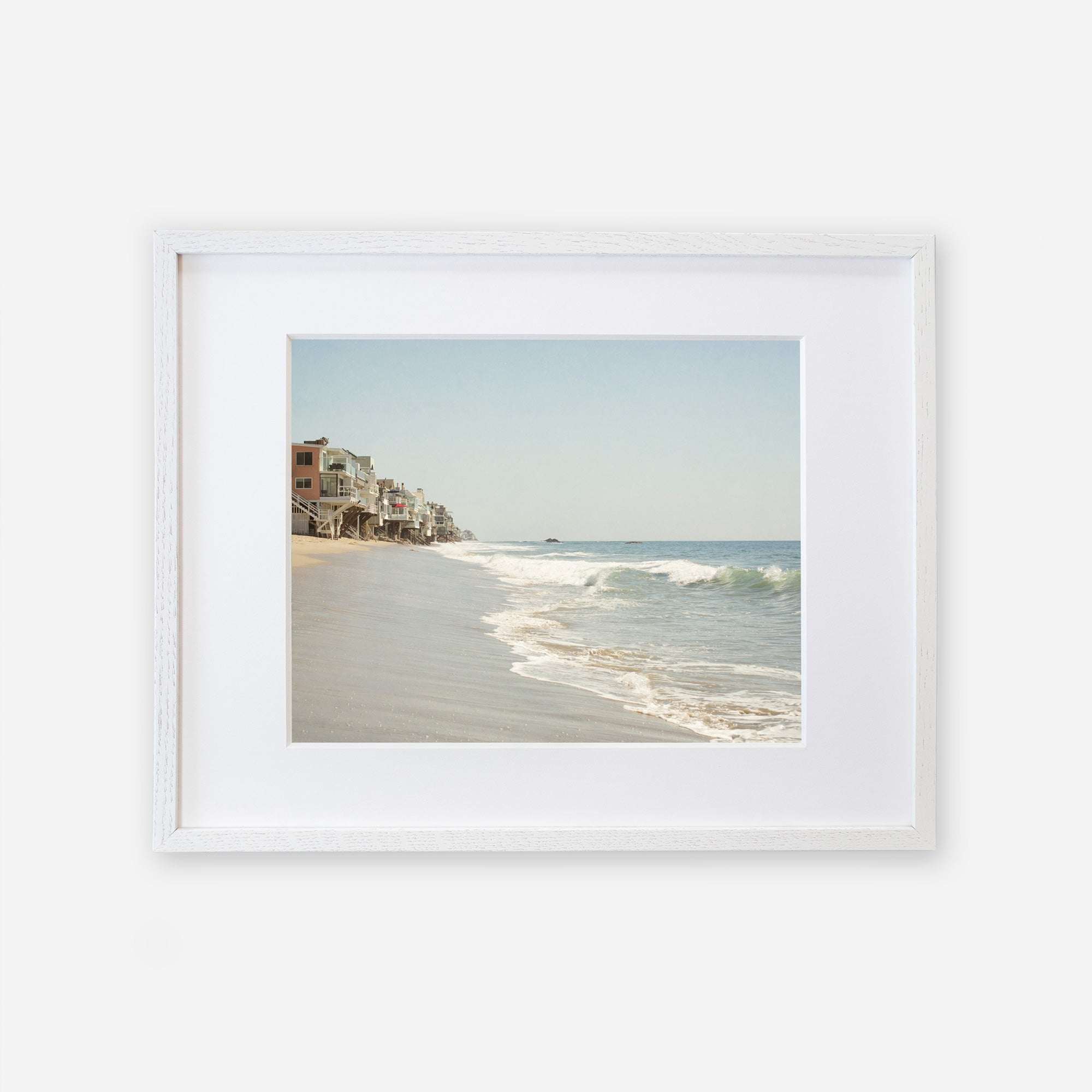 A framed Malibu Beach House Print, &#39;Ocean View&#39; of a serene Malibu coastline with waves gently lapping at the shore and a row of buildings extending along the coast under a clear sky by Offley Green.