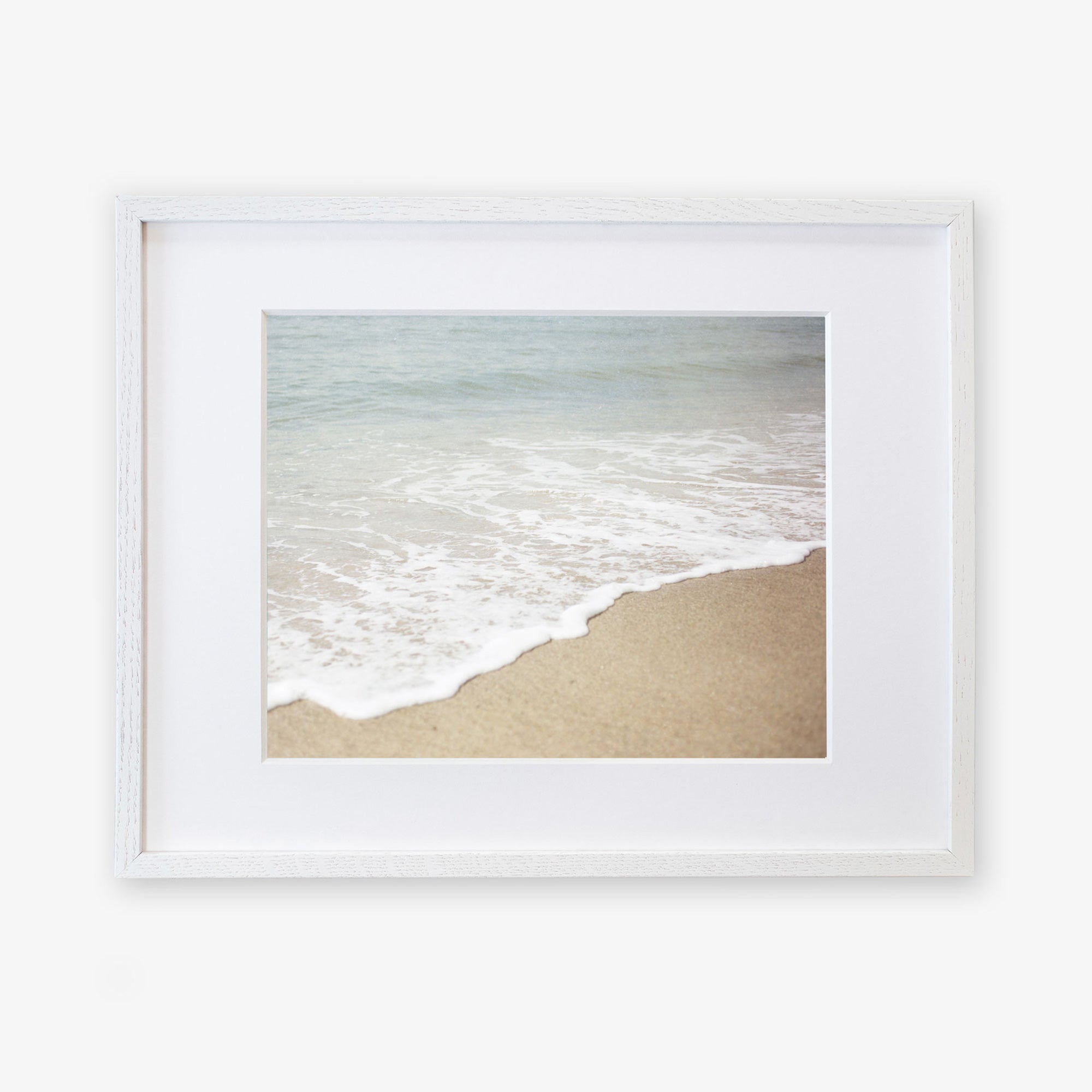 A framed photograph of a California beach with gentle waves lapping at the shore, set against a serene ocean backdrop. The frame is white and mounted on a white background featuring the Offley Green Beach Waves Print, &#39;Chasing Surf&#39;.