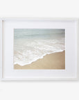 A framed photograph of a California beach with gentle waves lapping at the shore, set against a serene ocean backdrop. The frame is white and mounted on a white background featuring the Offley Green Beach Waves Print, 'Chasing Surf'.