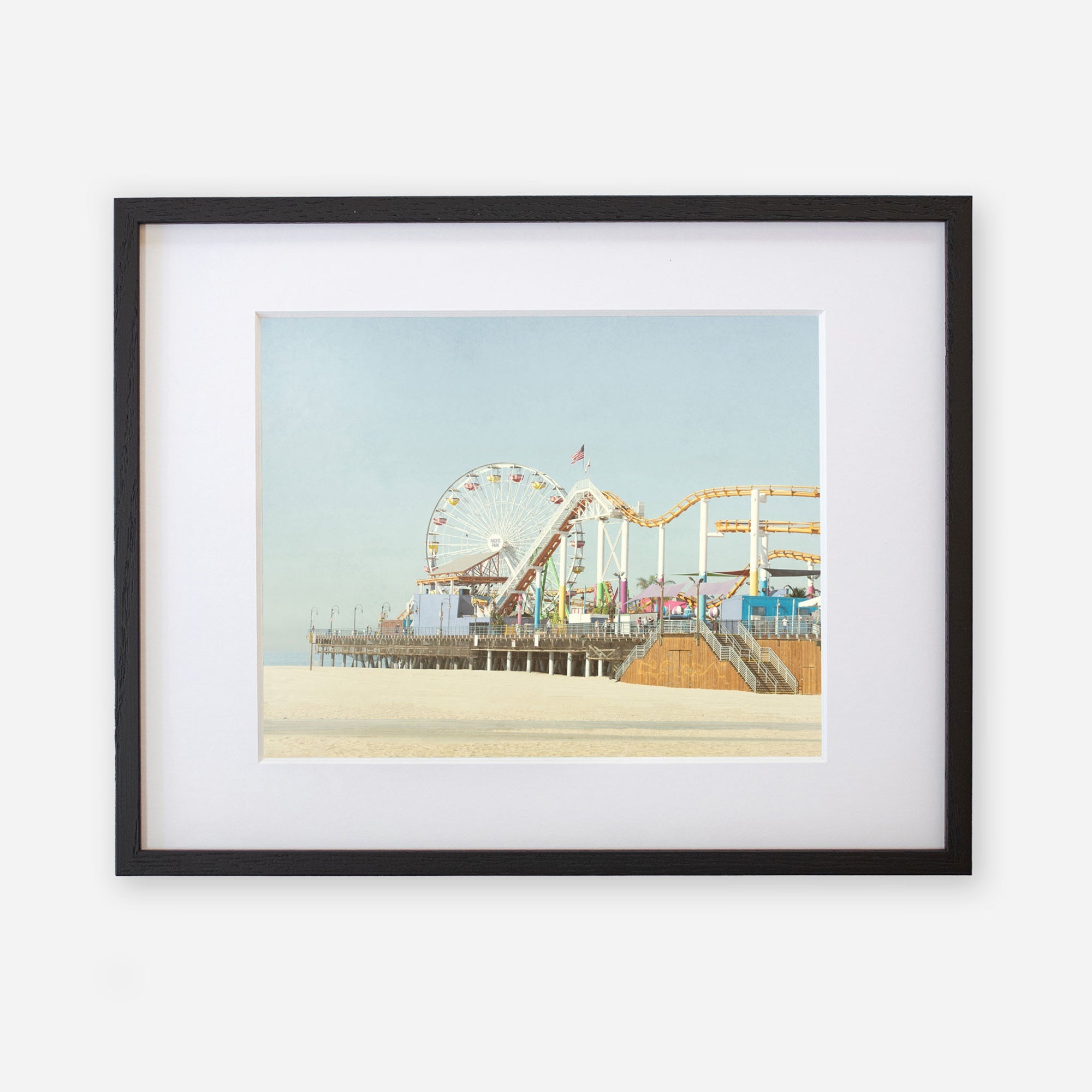 A framed Offley Green California Print, &#39;Santa Monica Pier&#39;, featuring a ferris wheel and roller coaster, displayed against a light blue sky.