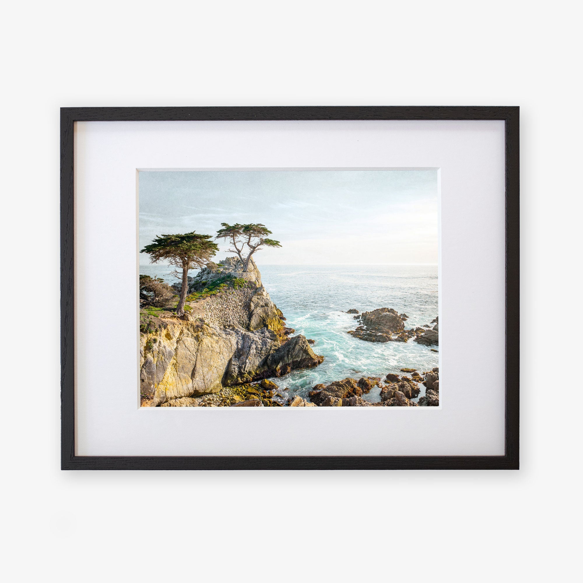 A framed painting of a rocky coastal scene with pine trees overlooking turbulent ocean waves, displayed against a white background on archival photographic paper. This is the Offley Green California Coastal Print, &#39;Lone Cypress&#39;.